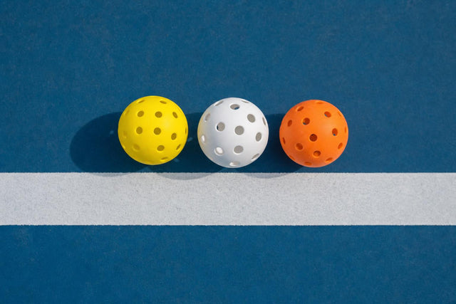 Una bola de pickleball amarilla de exterior, una blanca de interior y una naranja sobre una pista azul con una línea blanca