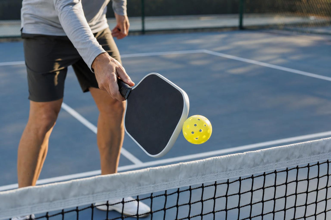 Close-up of a pickleball paddle gently touching a yellow ball next to the net, in the kitchen line