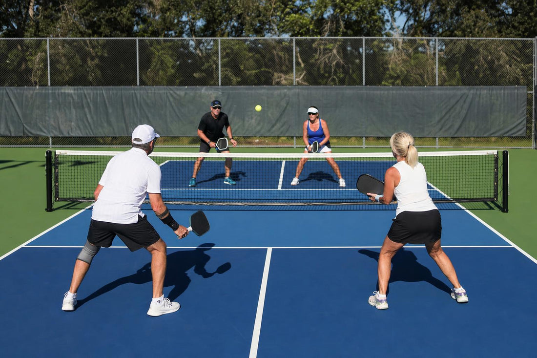 Cuatro jugadores en un partido de dobles de pickleball vistos desde el fondo de la pista, dos junto a la red y dos en el fondo