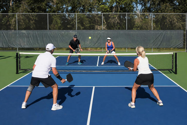Cuatro jugadores en un partido de dobles de pickleball vistos desde el fondo de la pista, dos junto a la red y dos en el fondo