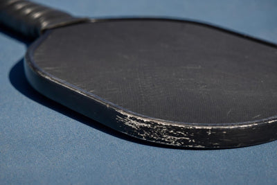 Detalle del canto y la cara de una pala de pickleball sobre una pista azul
