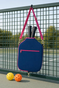 Navy blue CARRY cover hanging by its pink strap on a pickleball court fence, with two paddles inside and two balls on the ground