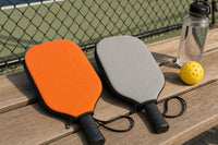 Two zip covers, one orange and one gray, on a bench next to a pickleball court with the paddles inside