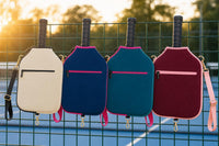 Four CARRY neoprene covers in cream, navy blue, teal and burgundy hanging from a pickleball court fence at sunset