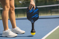 Close-up of a hand holding the paddle upside down by the head while the SCOOP ball pickup tool, on the end of the handle, holds a yellow ball at court level