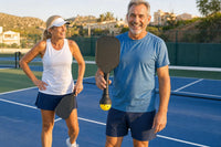 Two pickleball players smiling on court; one holds the paddle with the SCOOP and a ball held without having bent down